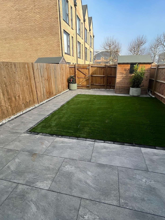 Backyard with gray patio stones and green grass, wooden fence, and buildings in the background.