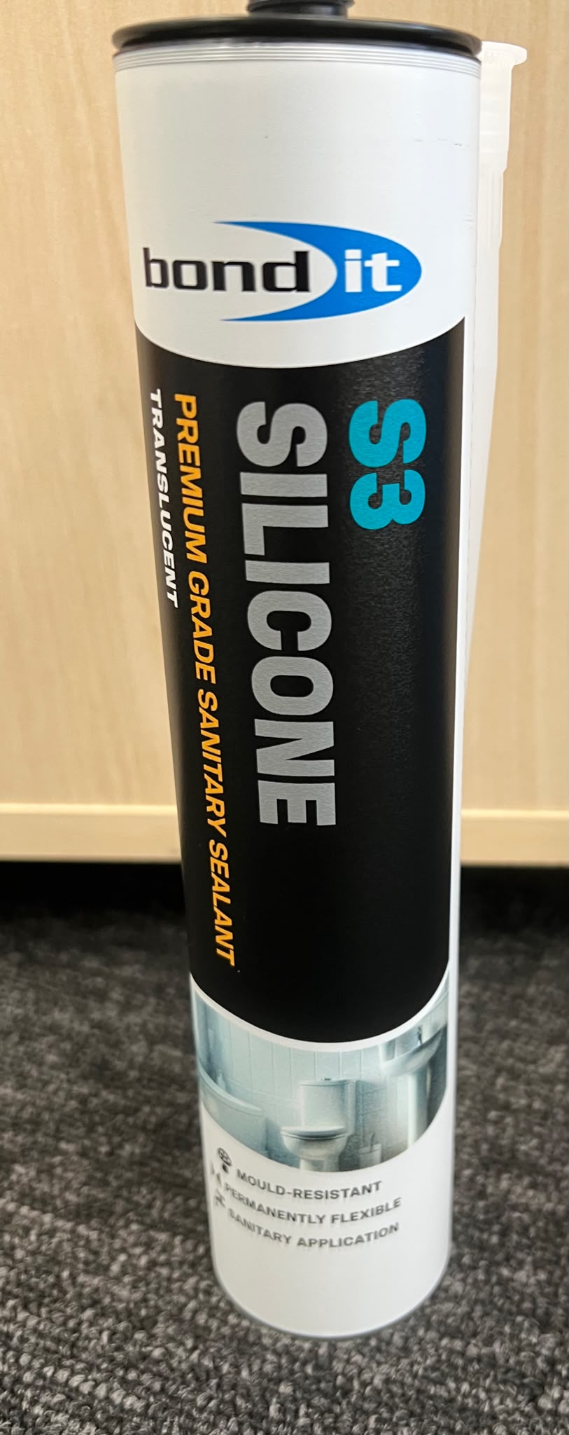 A tube of Bond It S3 Silicone Premium Grade Sanitary Sealant, featuring a white and black label with blue and orange text, standing on a gray carpet in front of a light wood panel wall.